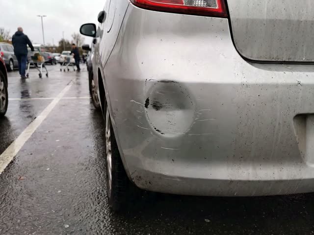 A still from the Claims Helper template: the rear bumper of a silver hatchback with a round dent and scuff marks, in a wet car park